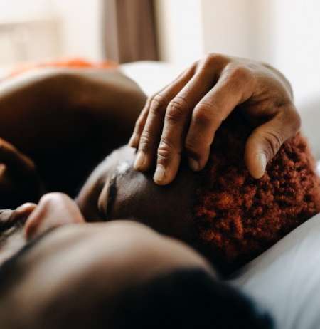 Close up on two dark-skinned persons' heads lying on a pillow while one person's hand gently cradles the other's head  