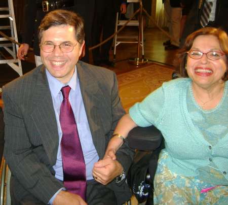 Two middle-aged white-skinned persons in wheelchairs wearing glasses and in formal attire, one male one female, smile broadly while holding hands.